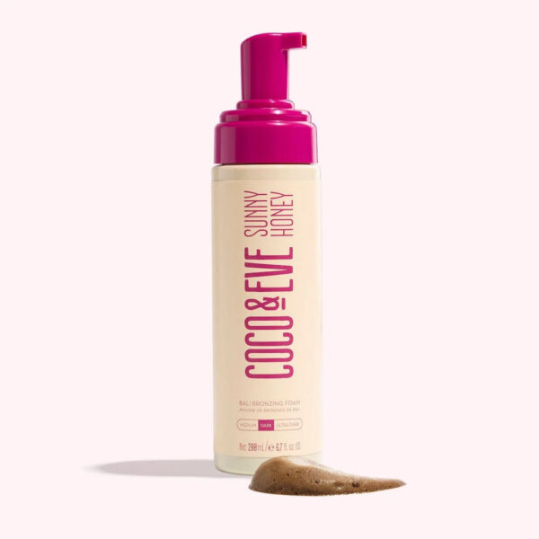 Close-up of Coco & Eve Sunny Honey self-tanner bottle against a backdrop of a tropical beach scene. The bottle features a vibrant label with tropical mango and guava fragrance, highlighting its organic ingredients and hydrating formula for a flawless, sun-kissed tan